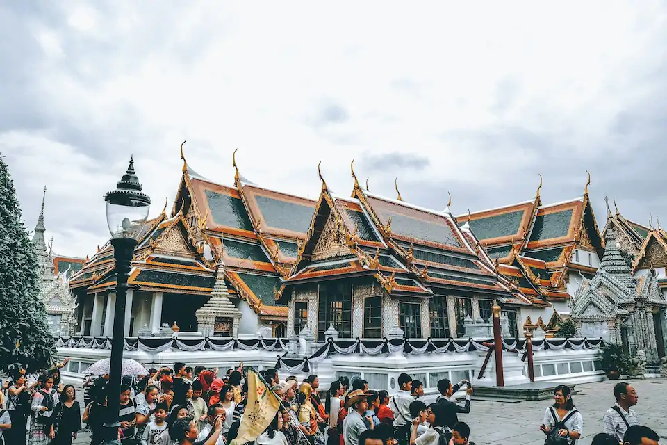 A large crowd of tourists and remote workers stands in front of ornate, traditional Thai temple buildings with intricate roofs and golden details under a cloudy sky.