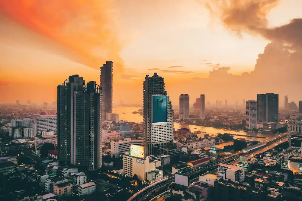 A city skyline at sunset, with tall buildings and skyscrapers alongside a river. The sky is ablaze with orange and yellow hues, casting a warm glow over Bangkok, one of the best cities in Thailand for remote workers.