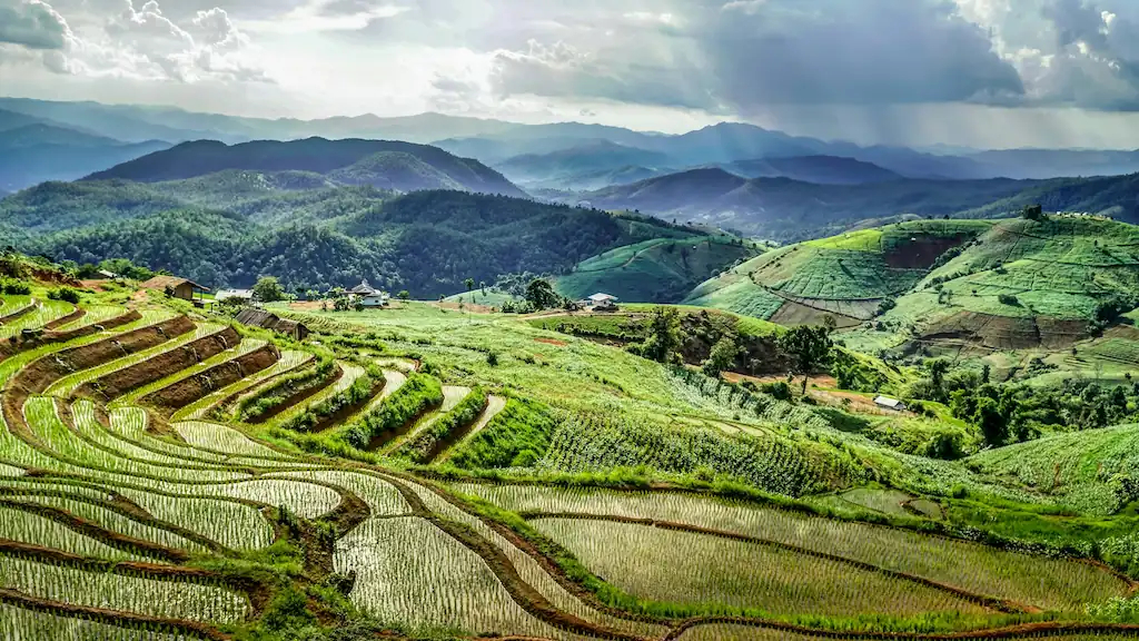 Terraced rice fields cover rolling green hills under a cloudy sky near Chiang Mai, with distant mountains and scattered small houses. Sunlight breaks through clouds, casting light on this vibrant landscape in one of the best cities in Thailand for remote workers.