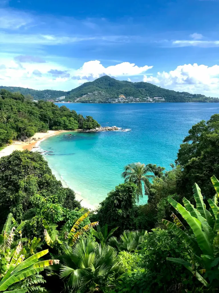 Tropical beach in Phuket with turquoise water, lush green palm trees, and foliage in the foreground; mountains and clouds in the background under a bright blue sky, one of the best cities in Thailand for remote workers.