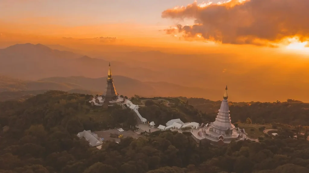 Thailand permanent residency pathway for remote workers - Aerial view of two pagodas on a lush green mountain at sunset, with golden light illuminating the sky and distant mountains.