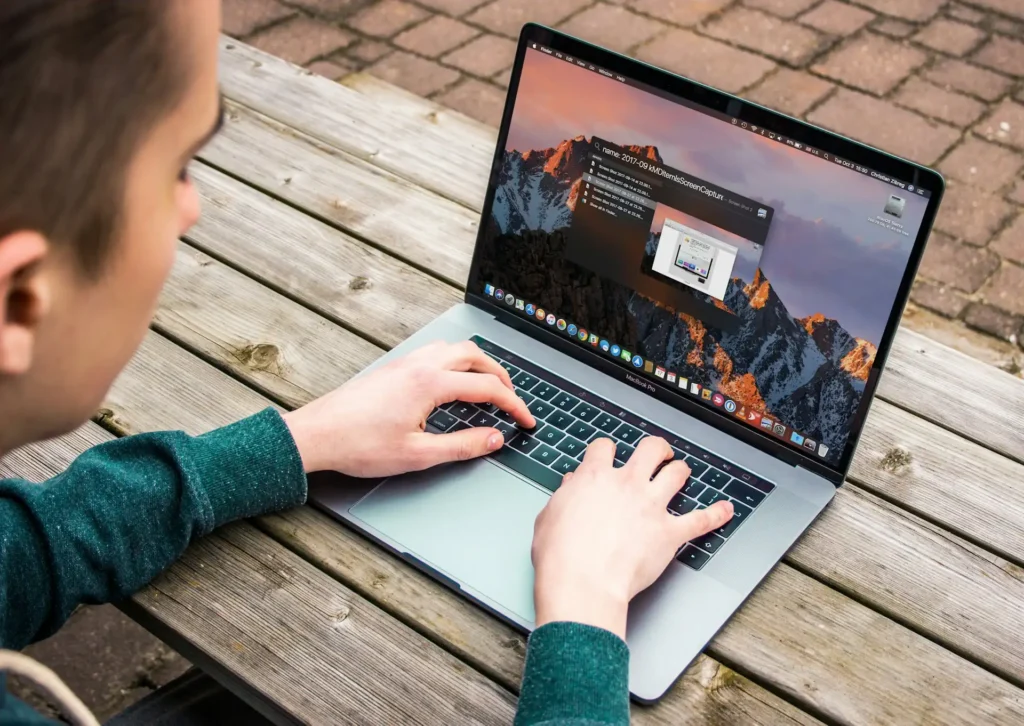 A person is using a laptop outdoors on a wooden table, researching business visa and work permit requirements for Thailand. The laptop screen displays an open application with a mountain landscape wallpaper as the user types with both hands.
