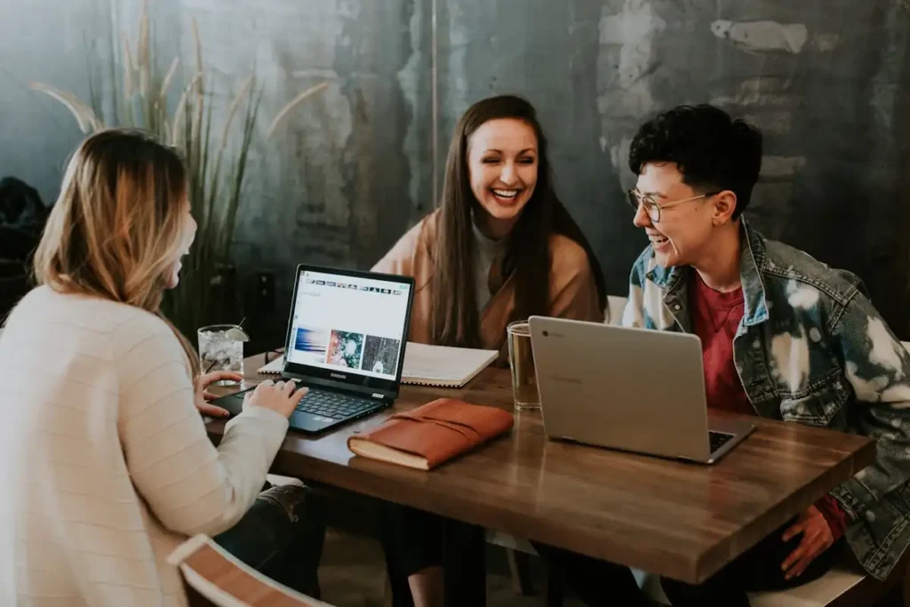 Three people sit at a wooden table with laptops, notebooks, and drinks, smiling and laughing as they discuss thailand work permit freelancer opportunities in a casual workspace with a dark, textured wall in the background.