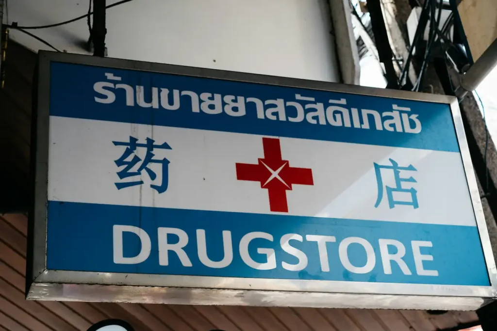 A sign for a drugstore featuring Thai language, a red medical cross in the center, and information about thai social security benefits, with DRUGSTORE in English at the bottom on a blue and white background.